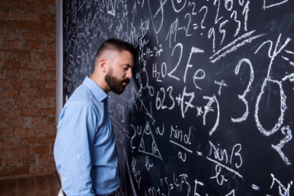 Man leaning his head against a chalkboard with math scrawled all over it